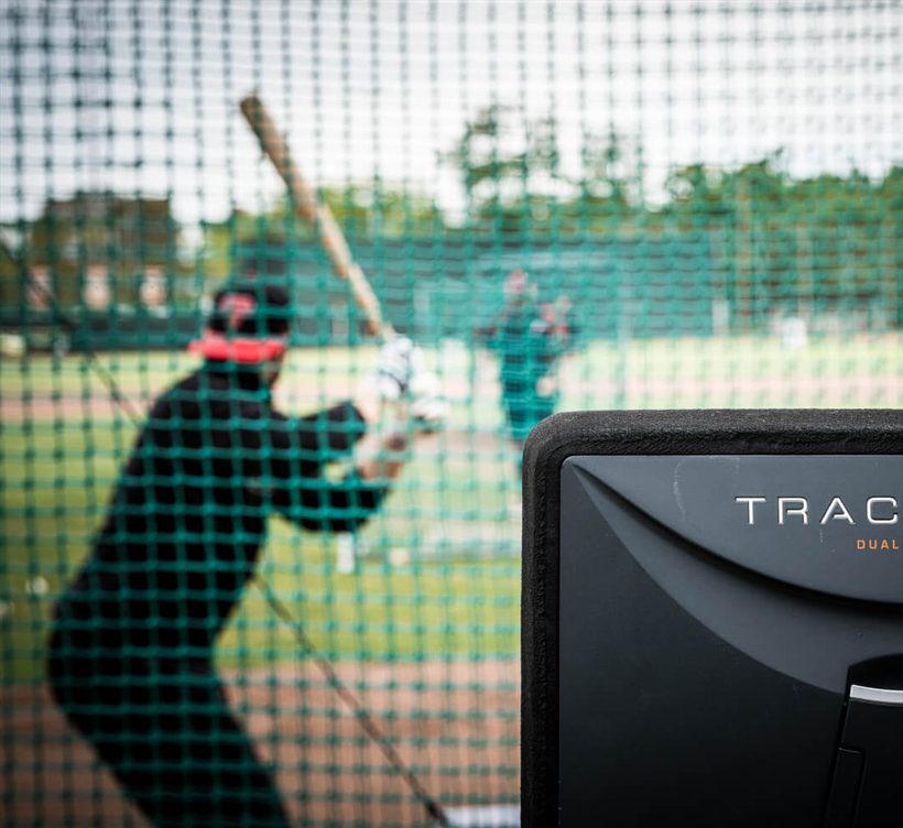 A hitter loads behind the cage net, out of focus, with the TrackMan unit in the foreground
