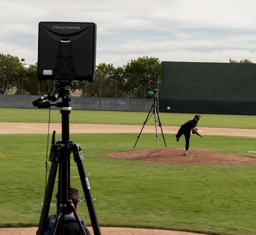 A TrackMan B1 on a tripod behind the mound as a pitcher follows through, the ball in flight