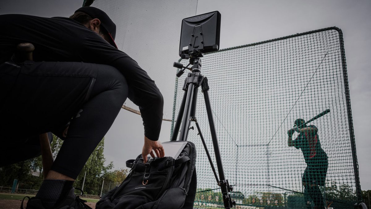 A TrackMan B1 unit on a tripod behind the cage net as a hitter loads, a coach crouched beside it
